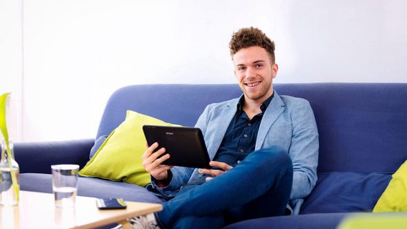 Junger Mann sitzt mit einem Tablet auf einem Sofa.
